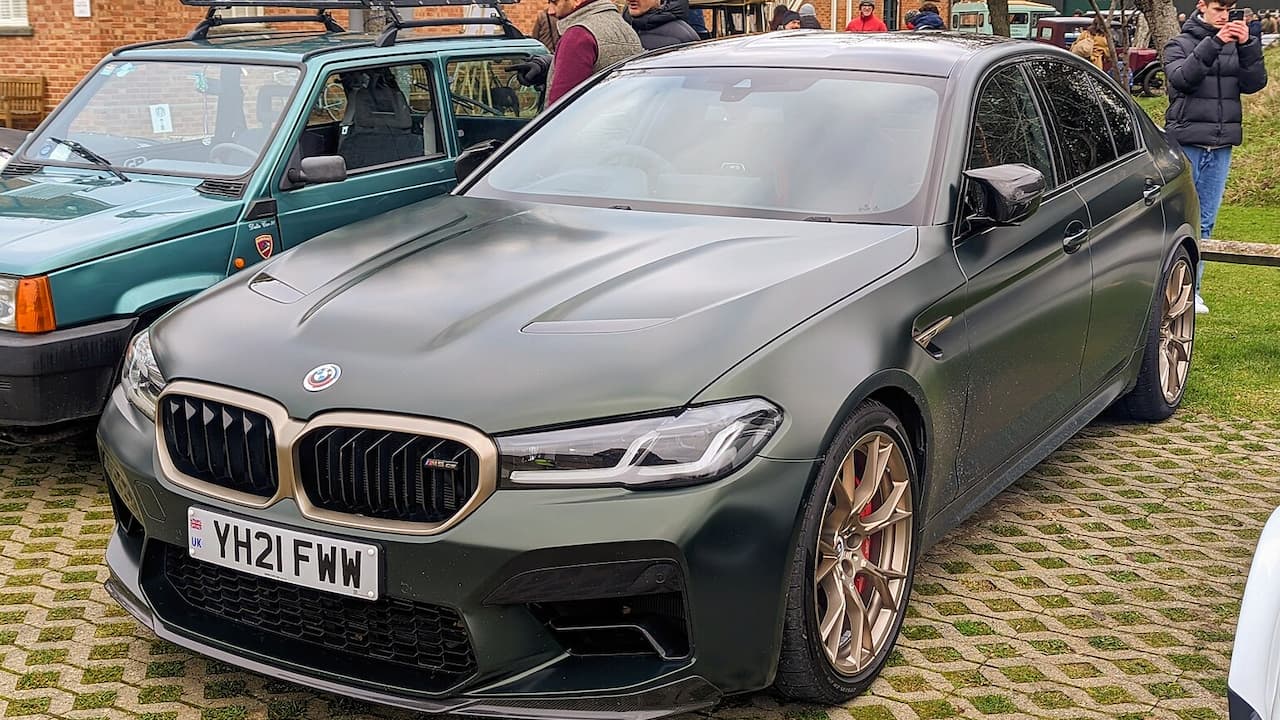 Dark green BMW M5 CS sedan with license plate "YH21 FWW" parked on cobblestones at car show with spectators