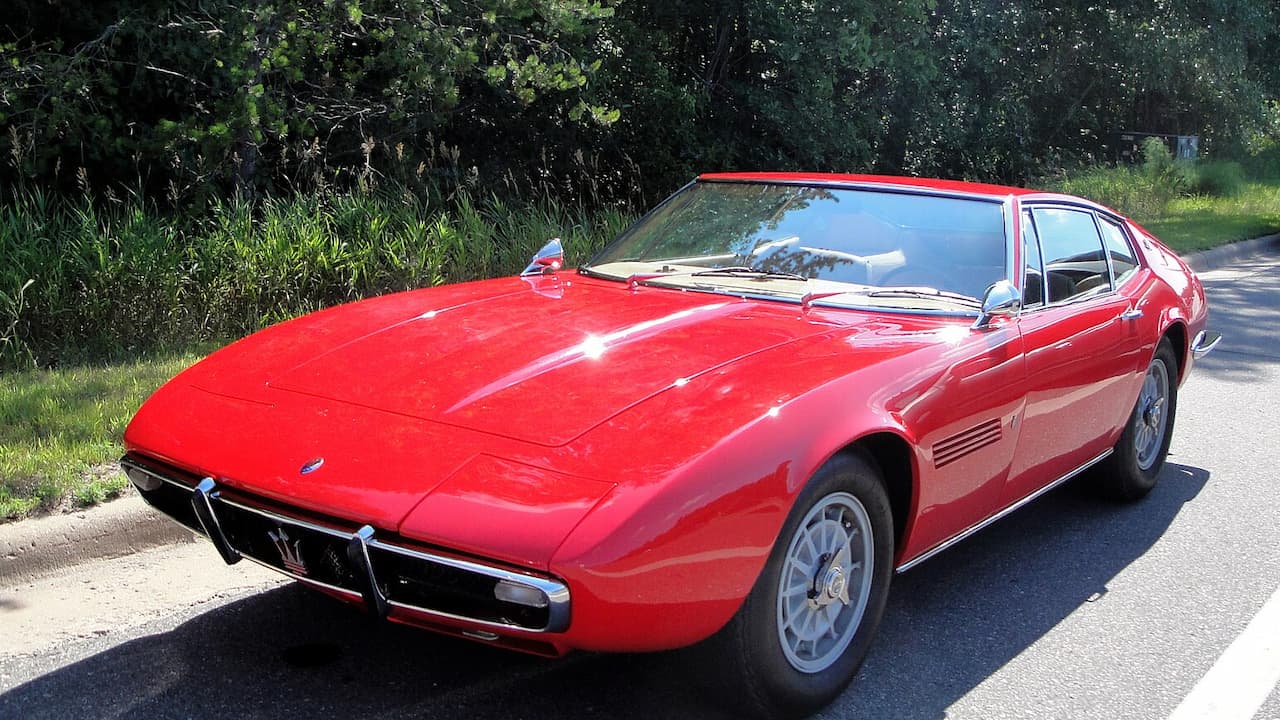 Red 1967 Maserati Ghibli with classic Italian design parked on asphalt road beside green vegetation