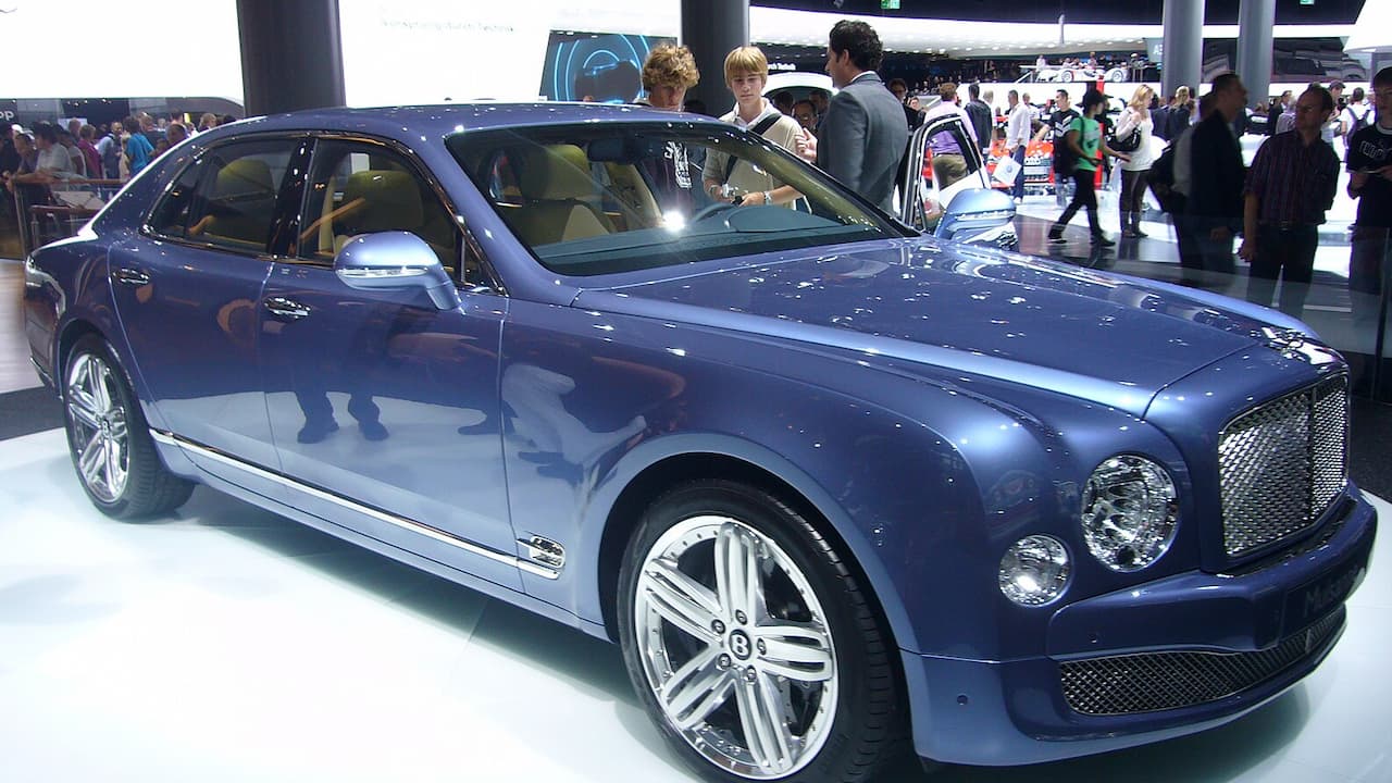 Blue Bentley Mulsanne displayed at auto show with chrome details, large wheels, and crowd of visitors viewing exhibits