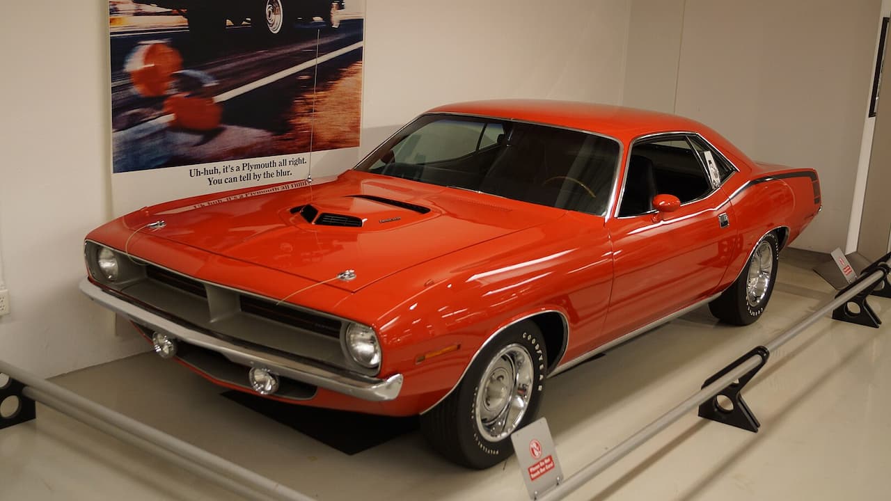 Orange 1970 Plymouth Hemi 'Cuda muscle car displayed in museum with vintage Plymouth advertisement poster behind