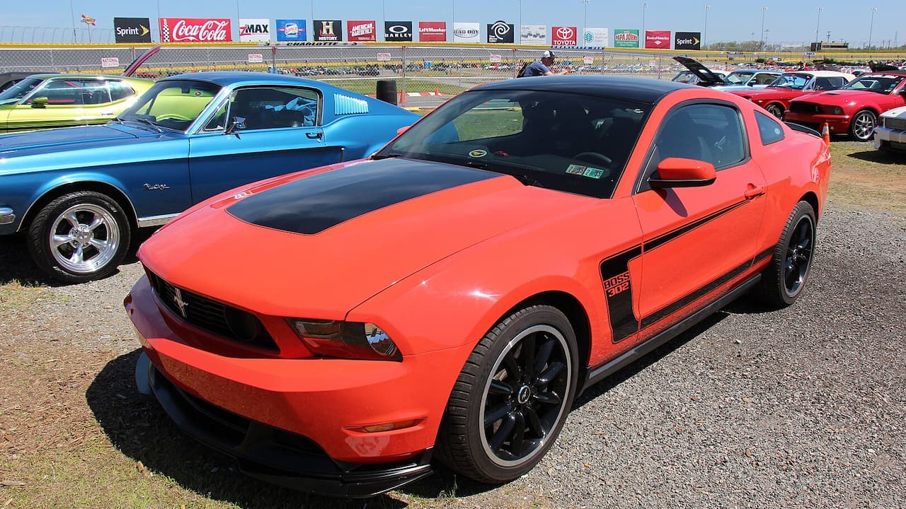 Orange 2012 Ford Mustang Boss 302 with black racing stripes and wheels parked at car show near racetrack