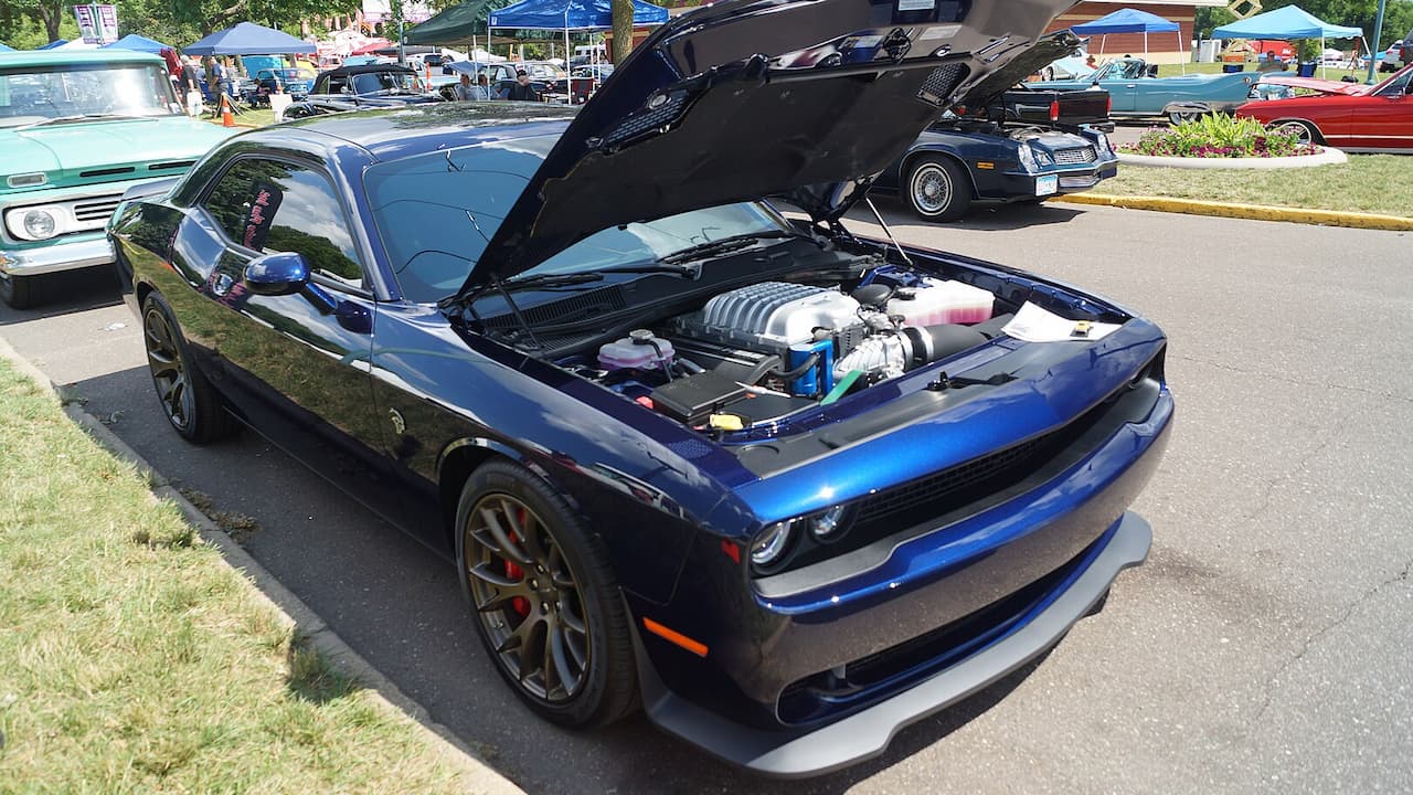 Blue 2015 Dodge Challenger Hellcat with hood open displaying supercharged engine at car show with other classic vehicles nearby