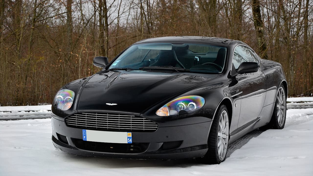 Black Aston Martin DB9 parked on snowy ground with bare winter trees in background