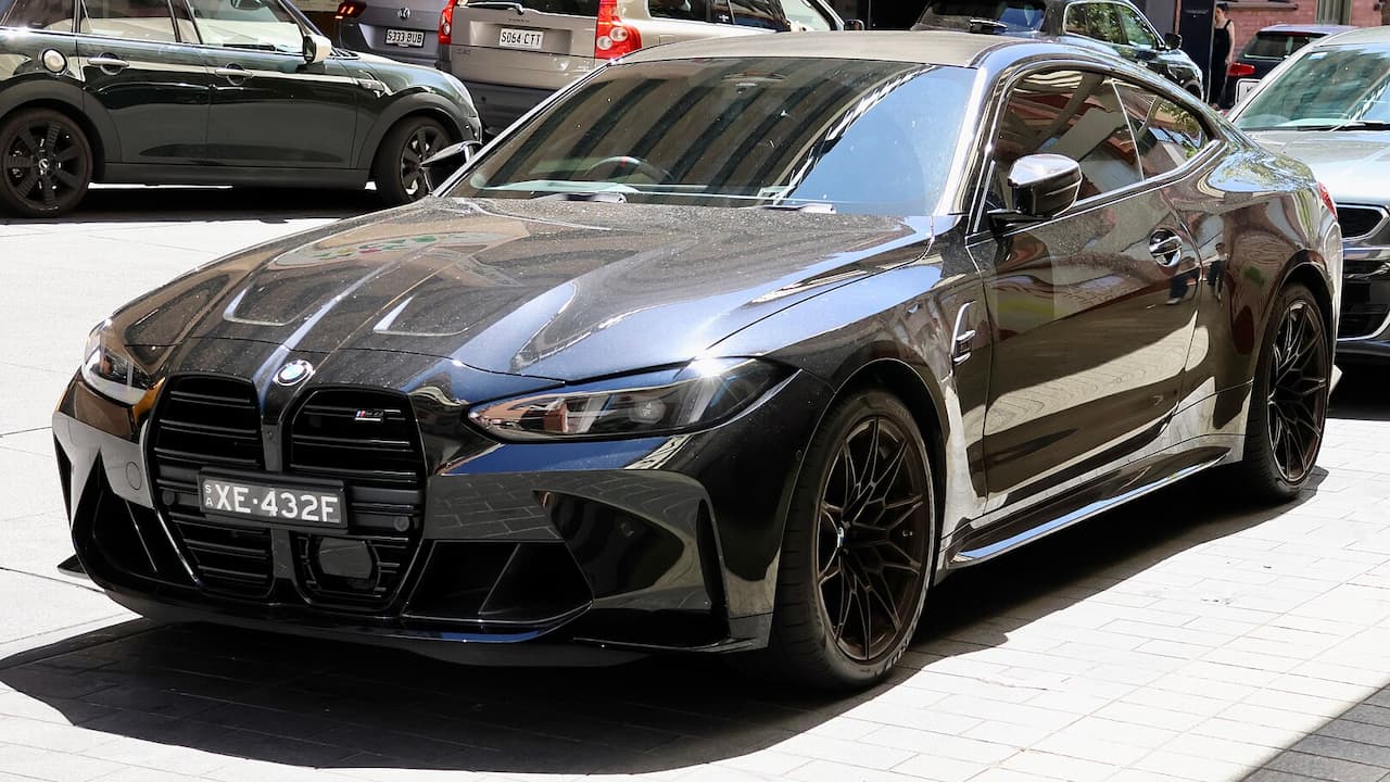 Black 2025 BMW M4 with distinctive kidney grille parked on street among other vehicles in urban setting