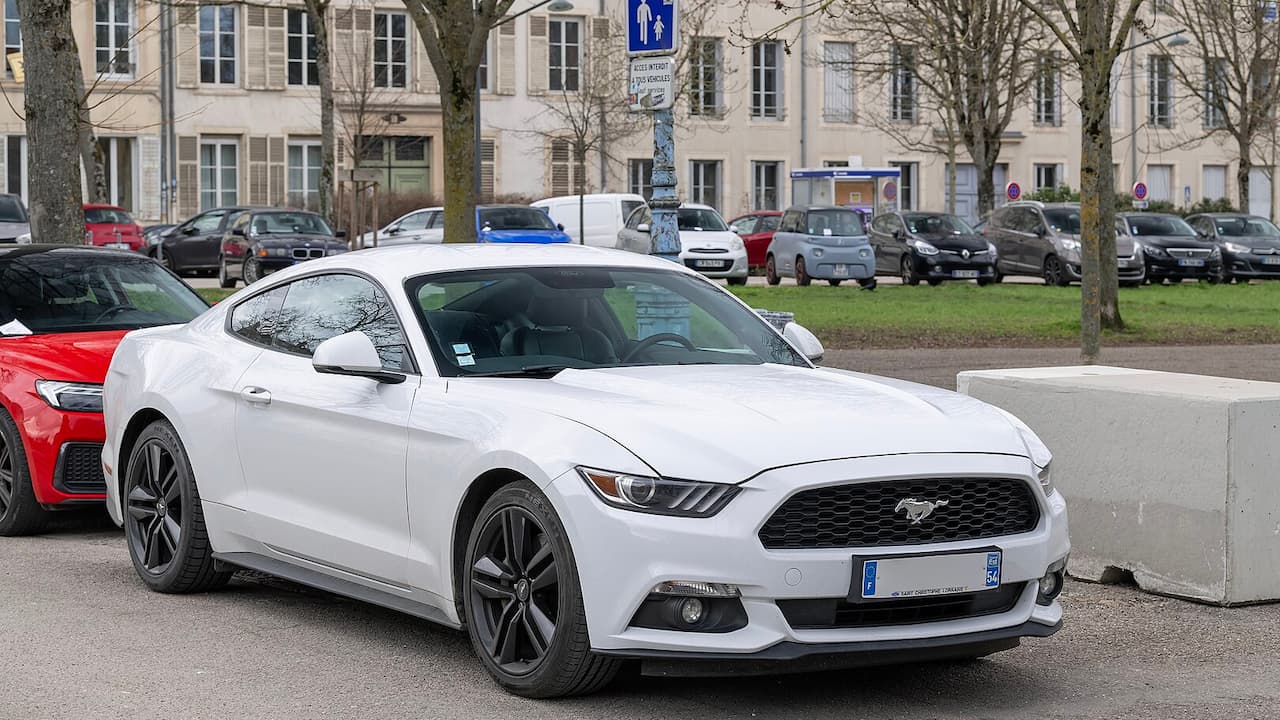 White 2025 Ford Mustang EcoBoost parked in European town square with historic buildings and other cars nearby