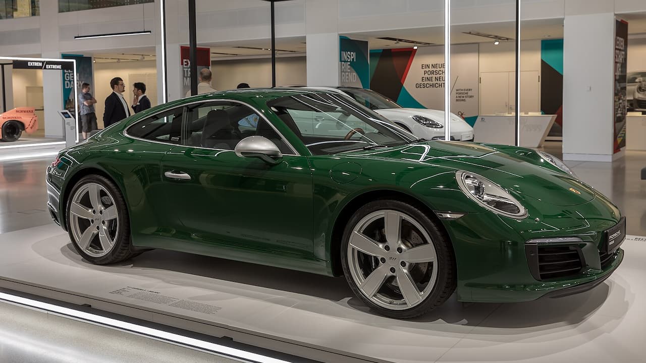 Green Porsche 911 displayed in modern showroom with glass barriers, visitors, and promotional banners in background