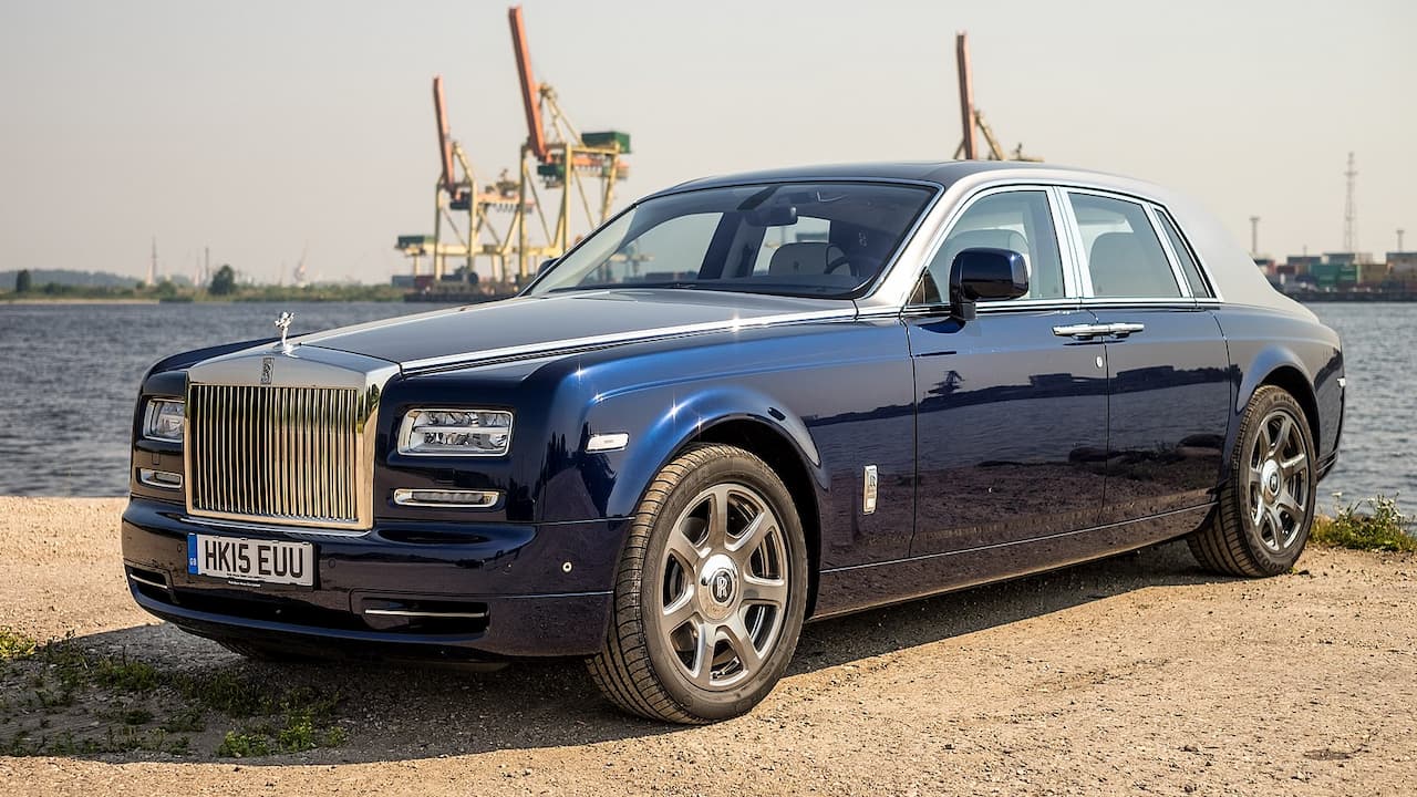 Dark blue Rolls-Royce Phantom with iconic vertical grille, Spirit of Ecstasy hood ornament, parked near industrial waterfront