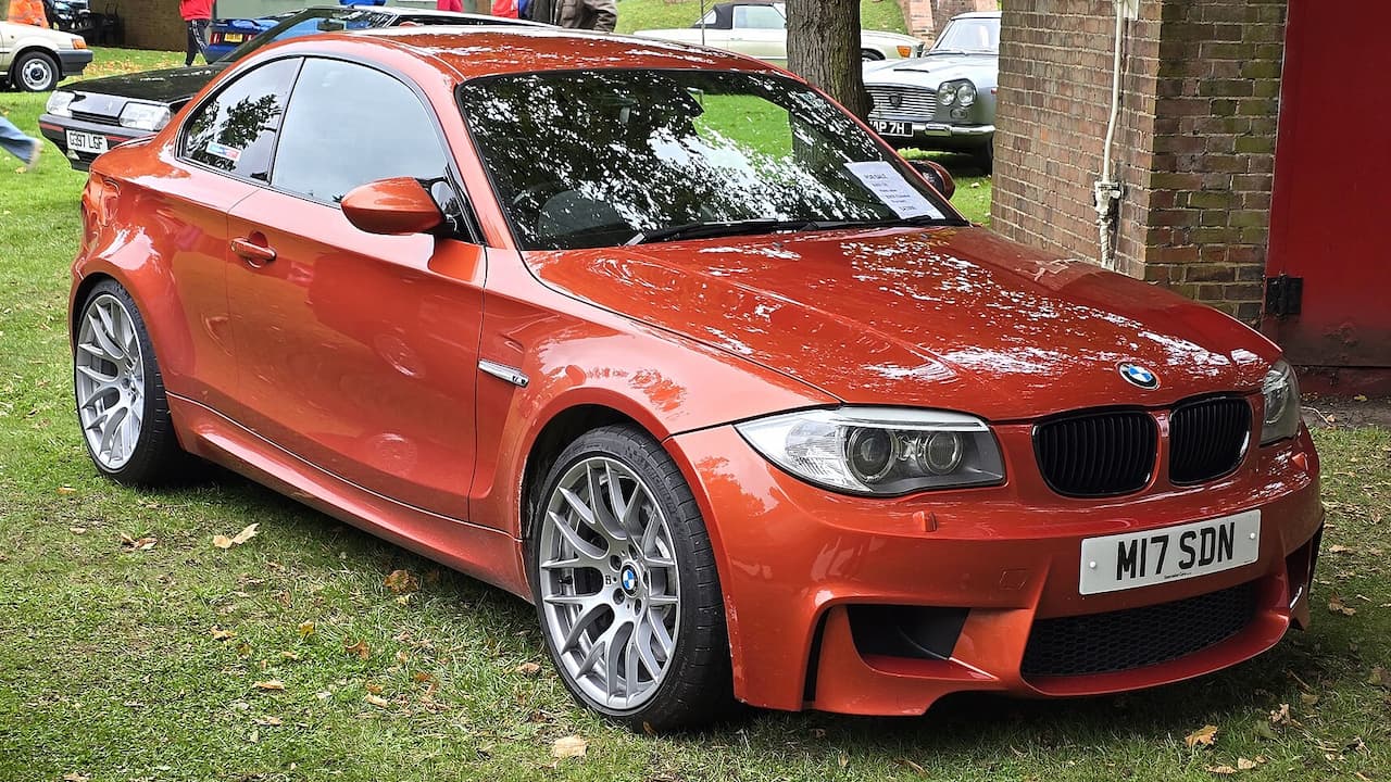 Orange BMW 1-Series M Coupe with silver alloy wheels parked on grass at car show with brick building background