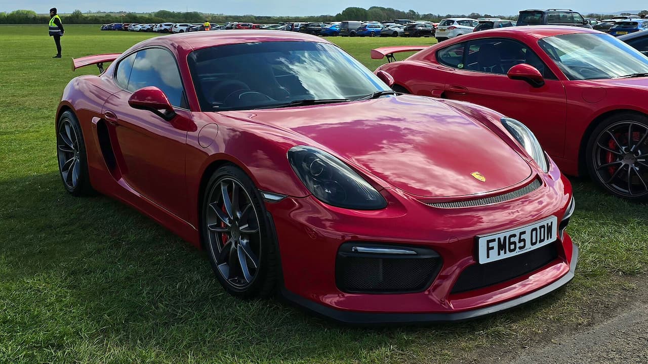 Red 2015 Porsche Cayman parked on grass field at car show with other vehicles and attendees visible