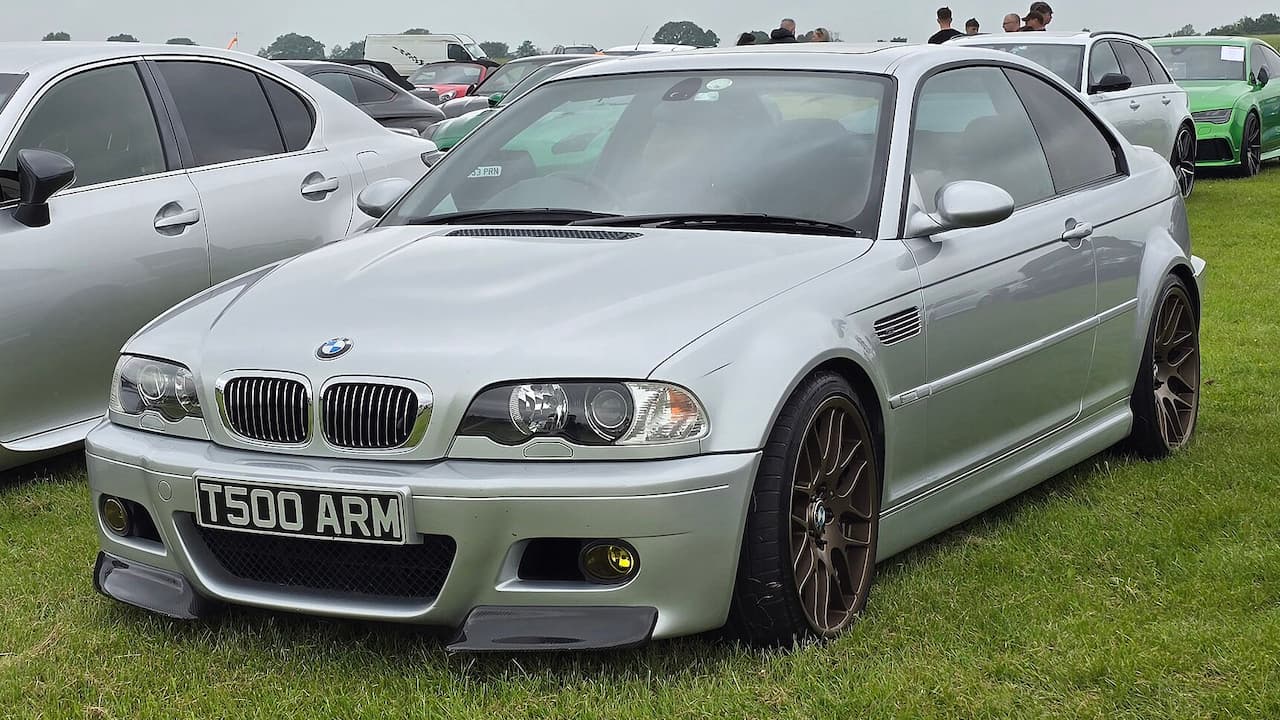 Silver BMW M3 with license plate T500 ARM parked on grass at outdoor car show with other vehicles nearby