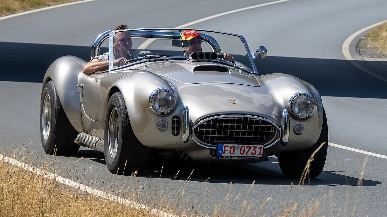 Silver 1969 Shelby Cobra convertible with two occupants driving on curved asphalt road with white lane markings and roadside vegetation