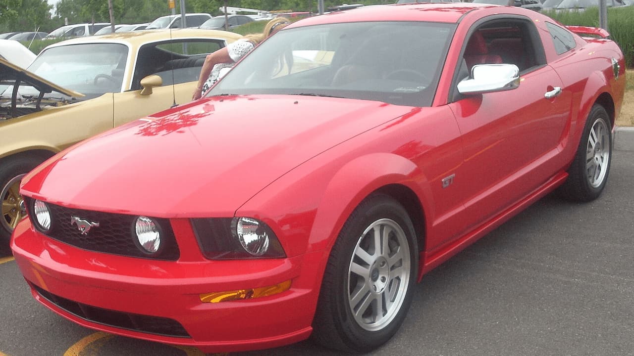 Red Ford Mustang GT (early 2000s) with chrome wheels and pony grille parked in lot with other classic cars nearby