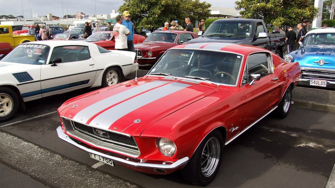 Red 1968 Ford Mustang Fastback with white racing stripes parked at outdoor classic car show with spectators