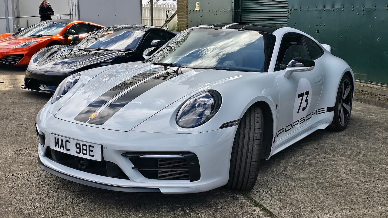 White Porsche 911 with racing stripes, number 73, and "PORSCHE" side decals parked among other supercars