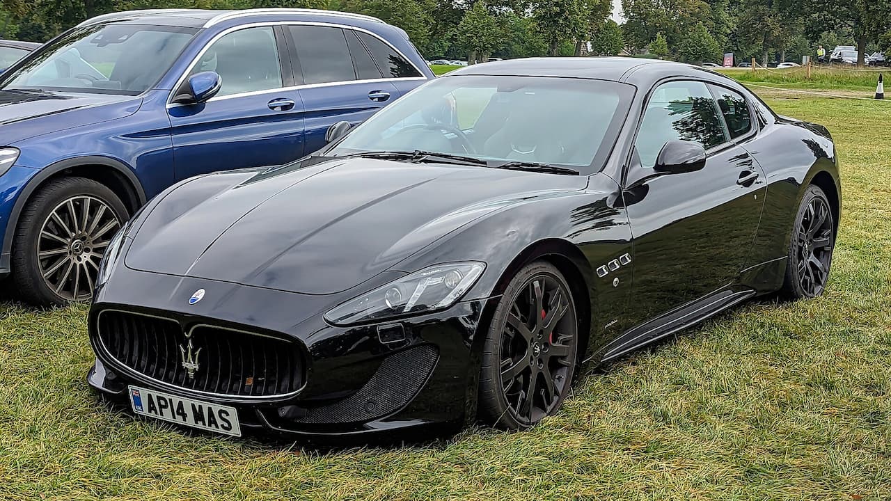 Black 2014 Maserati GranTurismo Sport with distinctive grille and license plate parked on grass with blue SUV nearby