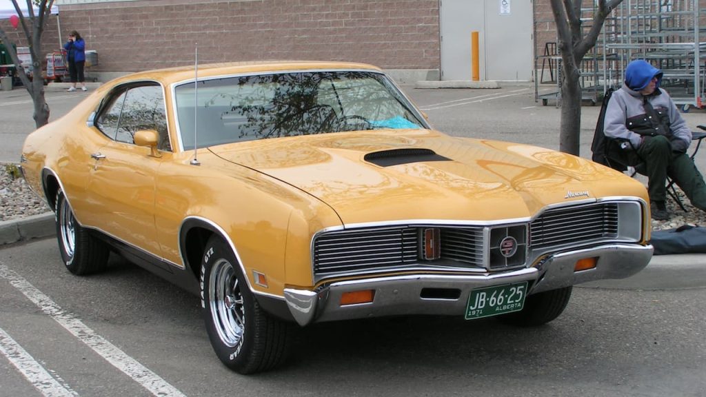 A gold 1971 Mercury Cyclone GT with chrome bumper and grille parked in a lot with Alberta license plate "JB-66-25". Trees frame the scene with people visible in background near a brick and metal commercial building