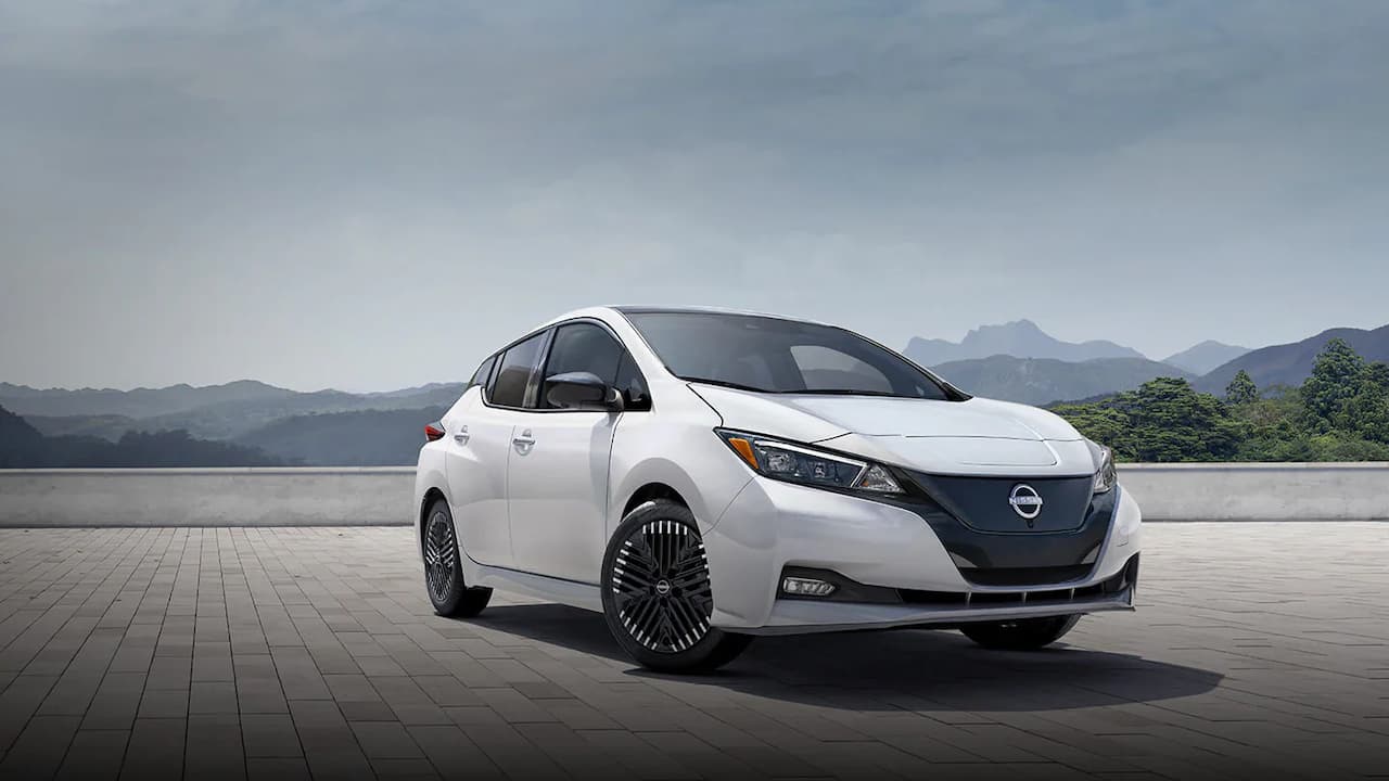 White 2025 Nissan Leaf electric hatchback with distinctive V-motion grille and turbine-style wheels on paved surface with mountain backdrop