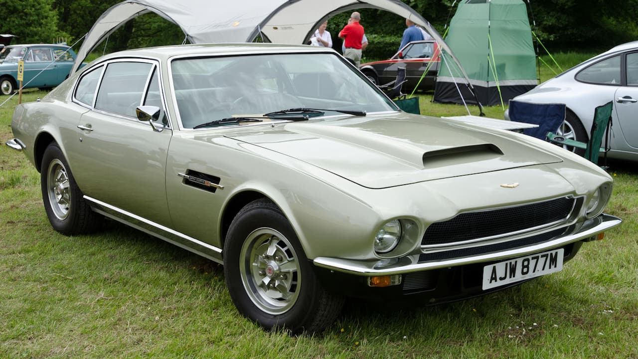 Silver 1973 Aston Martin V8 classic car parked on grass at outdoor car show with camping tents behind