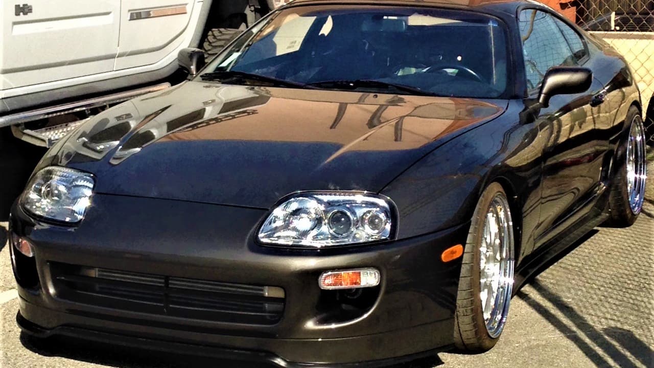 Black Toyota Supra (A80) sports car with chrome wheels and projector headlights parked on street near white vehicle