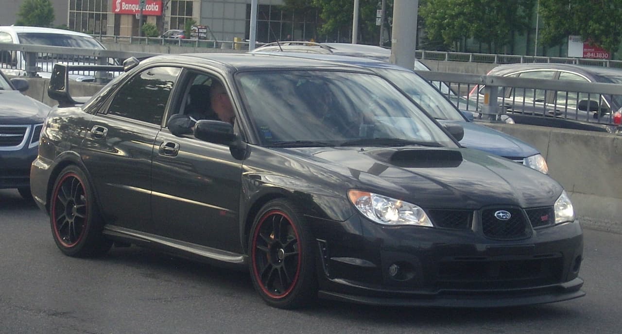 Black Subaru Impreza WRX STI with red wheel accents parked on city street with traffic and buildings