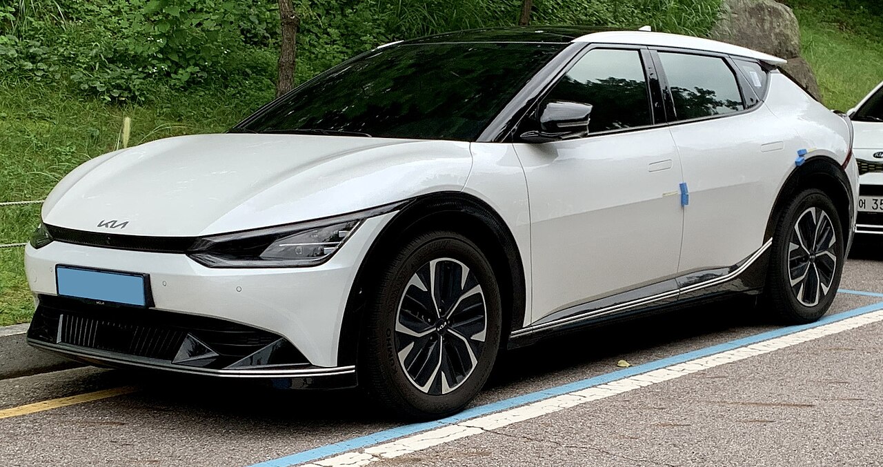White Kia EV6 electric crossover with sleek design and distinctive LED headlights parked in blue-marked parking space near greenery