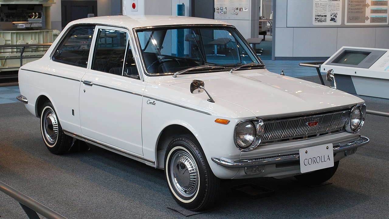 White 1966 Toyota Corolla two-door sedan on display in museum with "COROLLA" license plate and whitewall tires