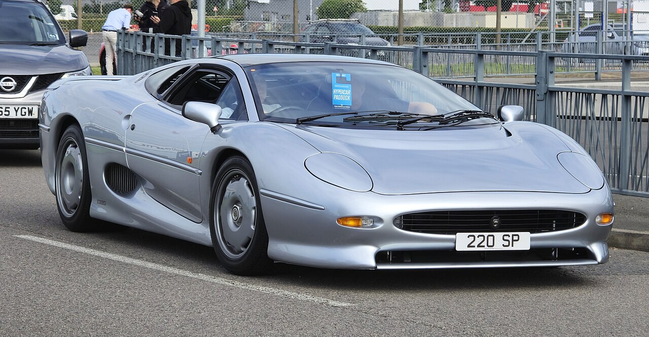 Silver 1993 Jaguar XJ220 supercar with distinctive oval headlights parked on street with metal barriers and spectators
