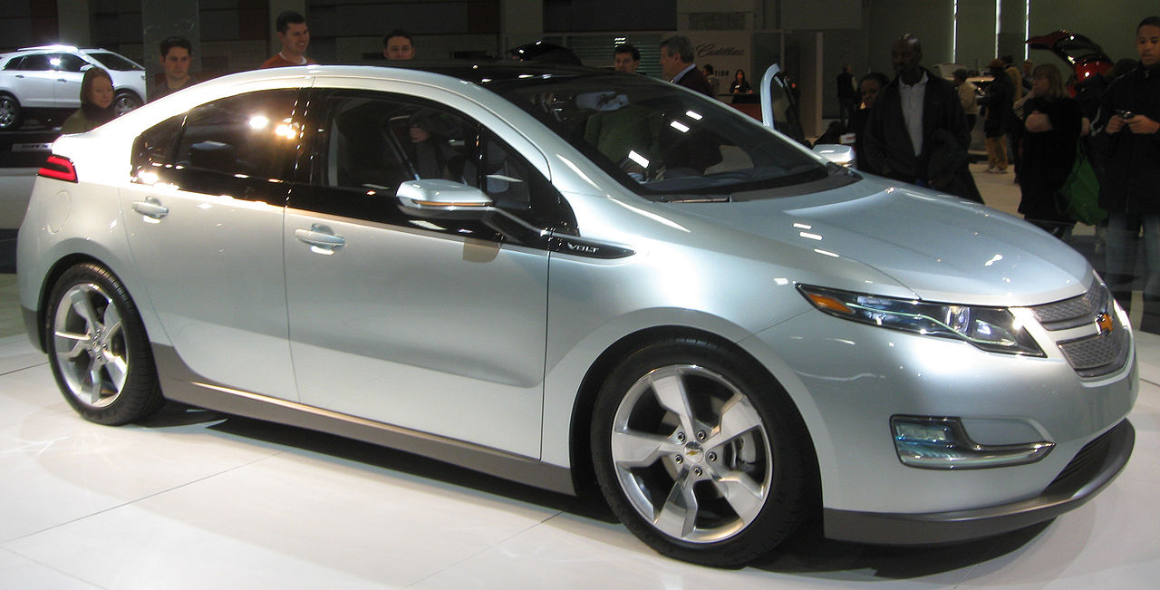 Silver 2010 Chevrolet Volt hybrid car displayed at auto show with crowd of onlookers and other vehicles visible
