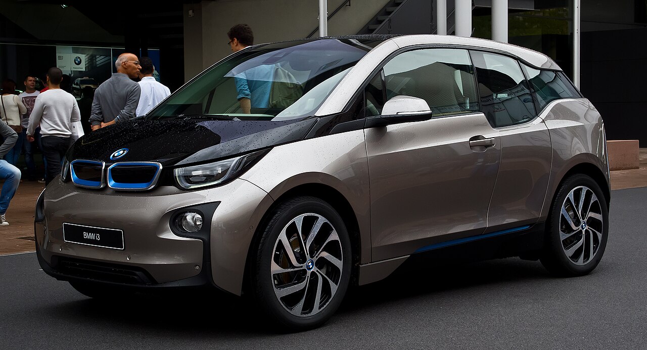 Bronze-colored 2013 BMW i3 electric car with distinctive design displayed at auto show with visitors observing nearby