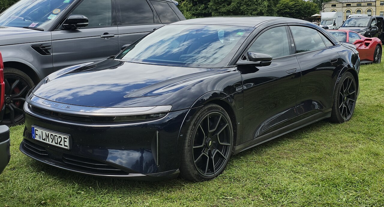 Dark blue Lucid Air Sapphire electric sedan parked on grass at car show with German license plate and other vehicles nearby