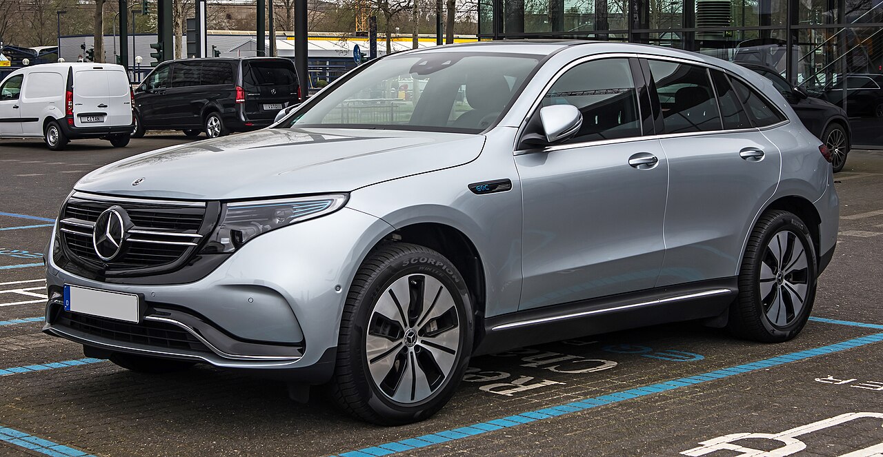 Silver Mercedes-Benz EQC electric SUV parked in blue-lined parking space with other vehicles and modern building visible