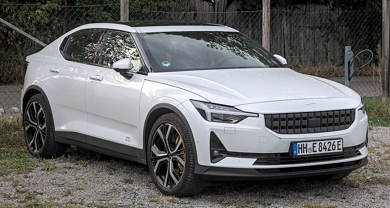 White Polestar 2 electric with minimalist design and dark alloy wheels parked on gravel near chain-link fence