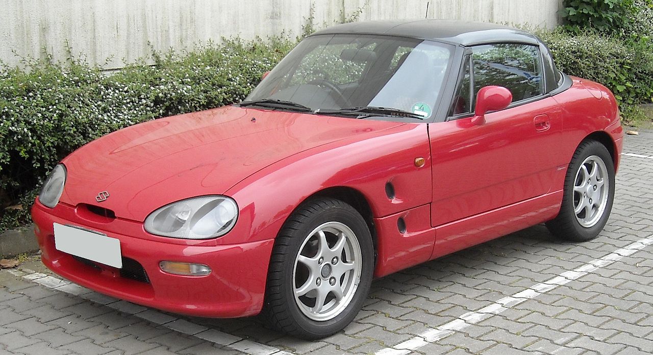 Red Suzuki Cappuccino with black soft top parked on cobblestone street beside concrete wall and green hedge