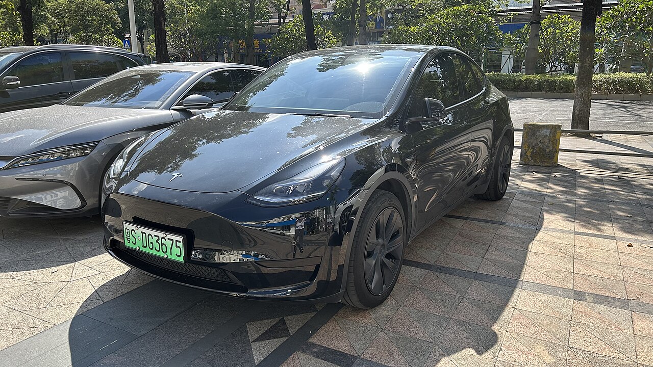 Black 2024 Tesla Model Y electric vehicle with green license plate parked on paved plaza with trees and buildings visible