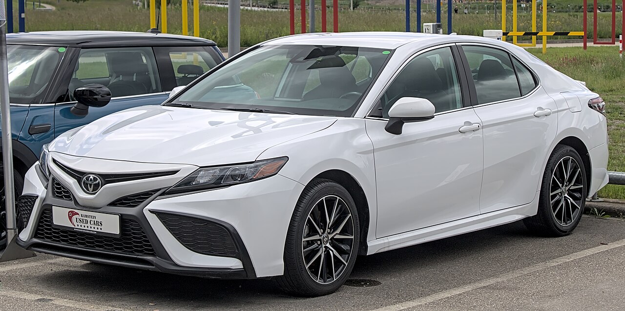 White Toyota Camry with sporty alloy wheels parked at used car dealership lot with colorful playground equipment visible