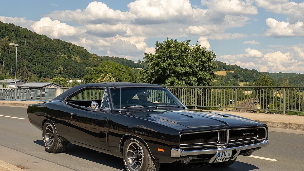 Black classic Dodge Charger muscle car parked on bridge with chrome bumper, rally wheels, and scenic hills behind