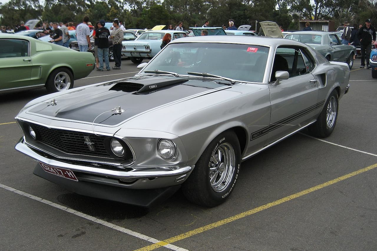 A silver Ford Mustang Mach 1 with black hood stripes and chrome wheels parked at an outdoor car show