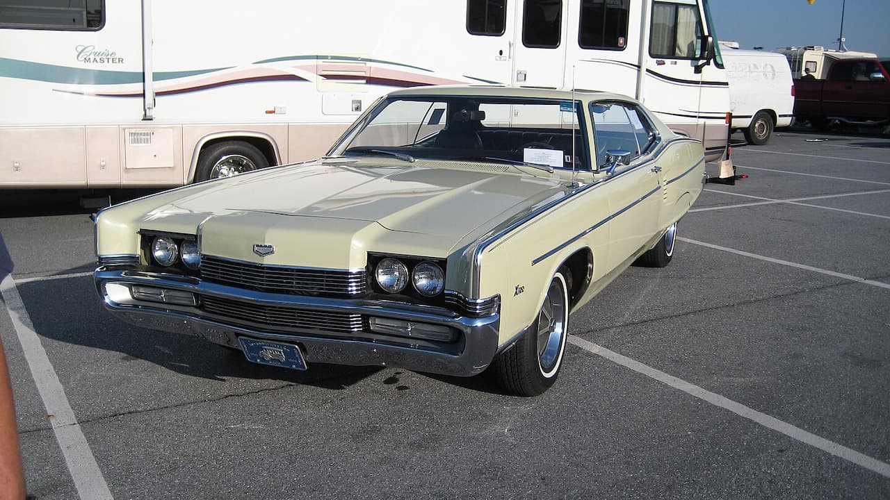 Classic 1969 Mercury Marauder muscle car with cream paint and blue racing stripes parked in lot near RVs