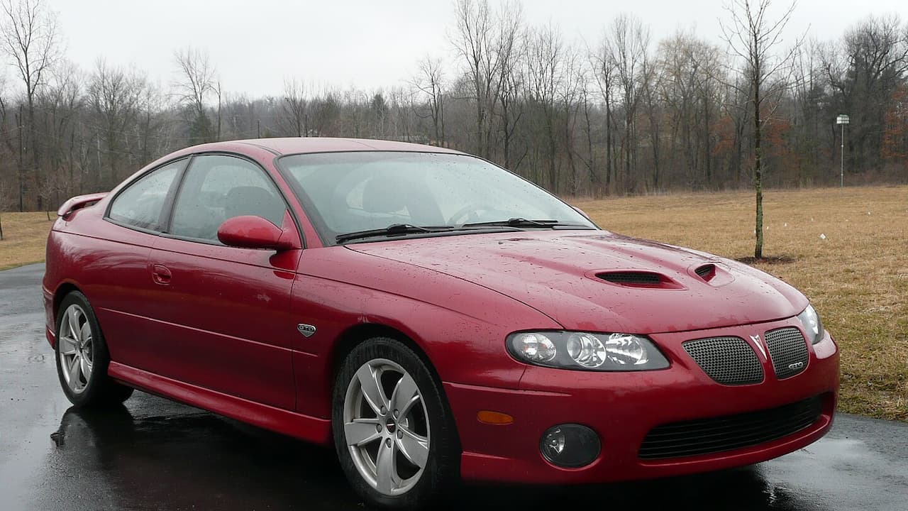 Red 2006 Pontiac GTO muscle car with hood scoops parked on wet pavement in rural field setting