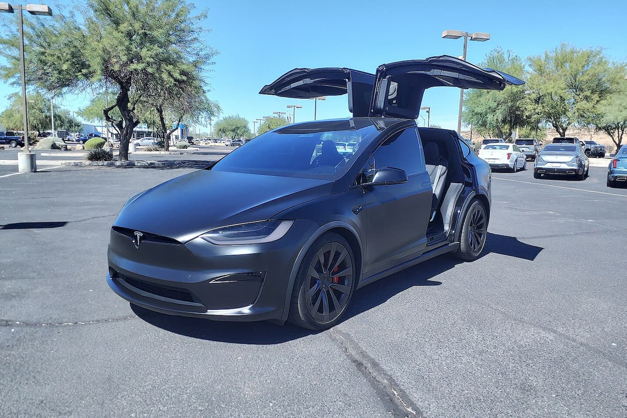 Dark gray Tesla Model X Plaid with distinctive falcon wing doors open, parked in sunny parking lot with other vehicles