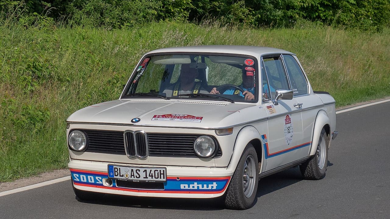 White BMW 2002 with racing livery and German license plate driving on road with grassy background