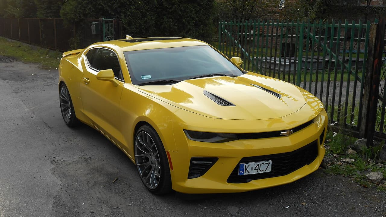 Yellow Chevrolet Camaro SS sports car parked on asphalt near green metal fence with hood vents and alloy wheels