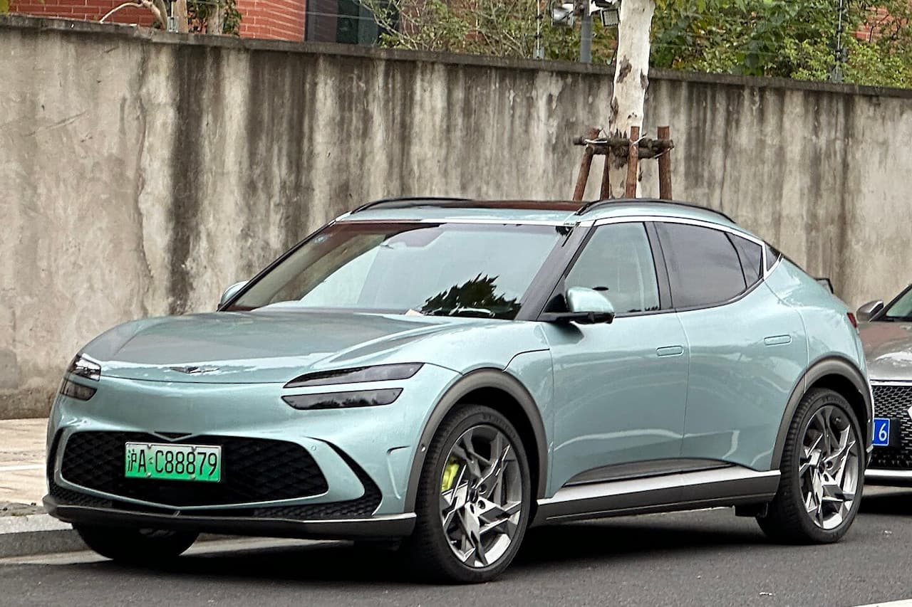 Light green Genesis GV60 electric SUV with distinctive LED headlights and modern design parked on street beside concrete wall