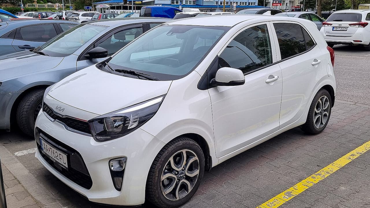 White Kia Picanto parked in busy parking lot with distinctive tiger-nose grille and stylish alloy wheels among other vehicles