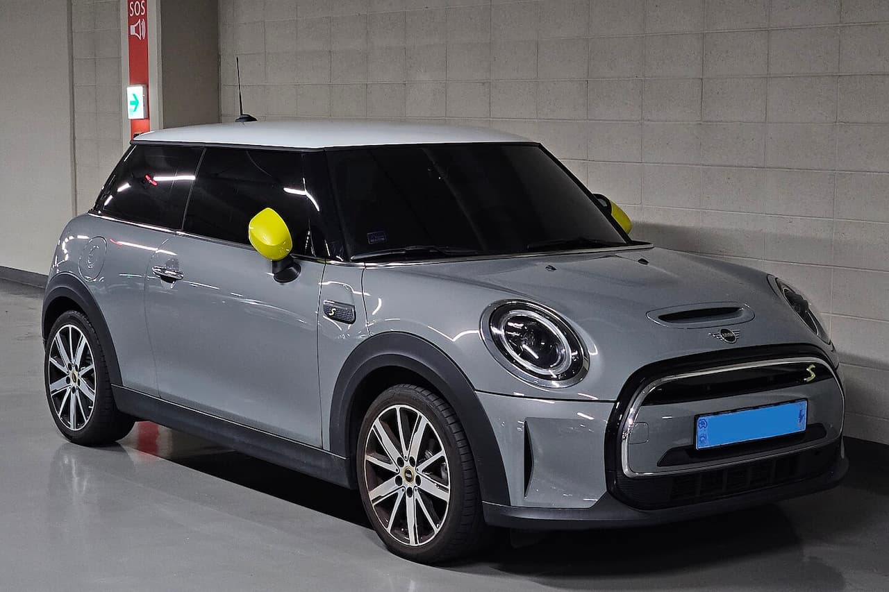 Silver Mini Electric Cooper SE with yellow mirror caps and closed grille parked in concrete garage near SOS phone