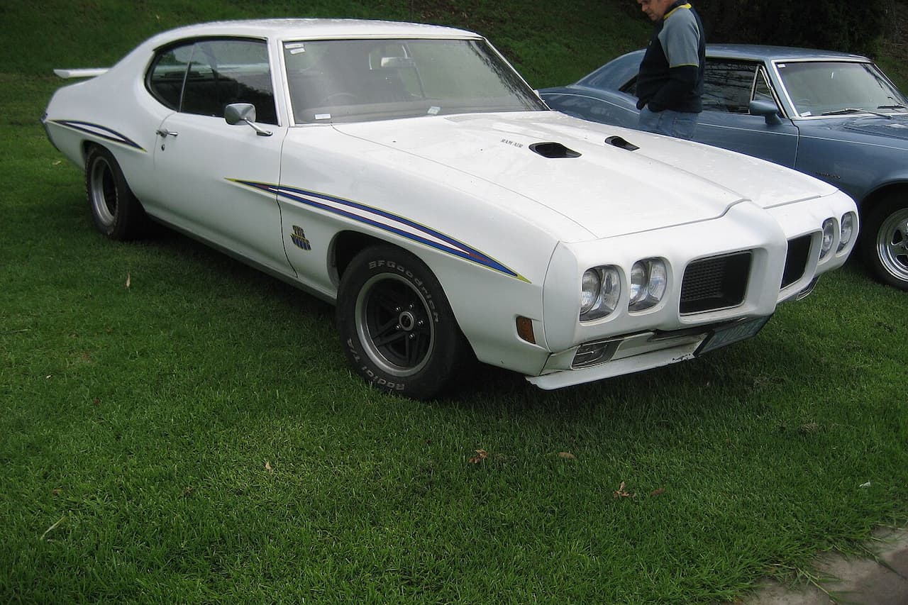 White vintage muscle car, a 1970 Pontiac GTO Judge, parked on green grass. Sleek design with blue racing stripes, muscular body, and black wheels. A person stands near another car in the background