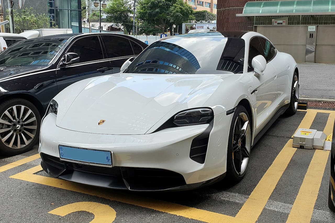 White Porsche Taycan Turbo S electric sports car parked in yellow-marked parking space with black sedan nearby in urban setting