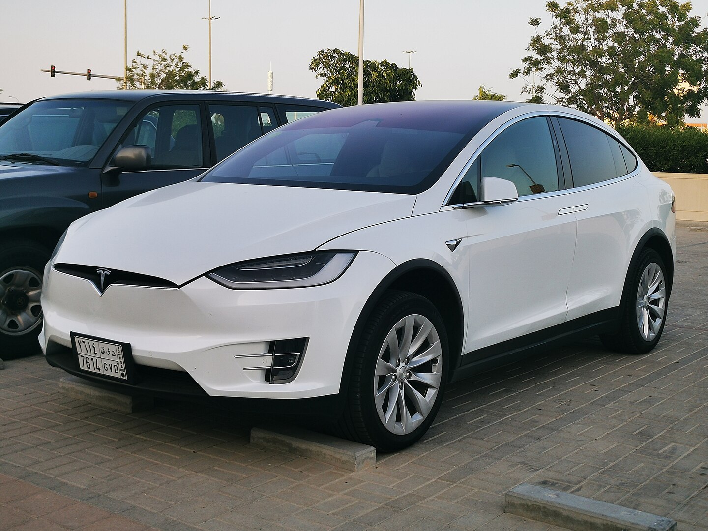 White Tesla Model X SUV parked on brick pavement next to dark vehicle, featuring distinctive falcon-wing doors and Tesla logo