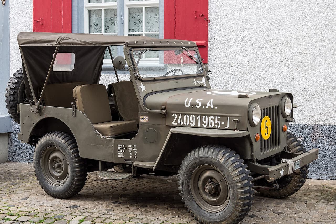 Olive green military Jeep with U.S.A. markings, canvas top, and knobby tires parked on cobblestone street near building