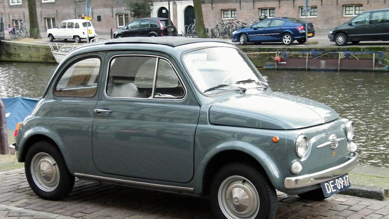 Fiat 500 with Dutch license plate parked on cobblestones beside canal with buildings behind